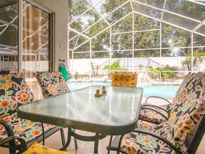 A glass-top table with chairs, a pool, and a view of trees and a white fence.