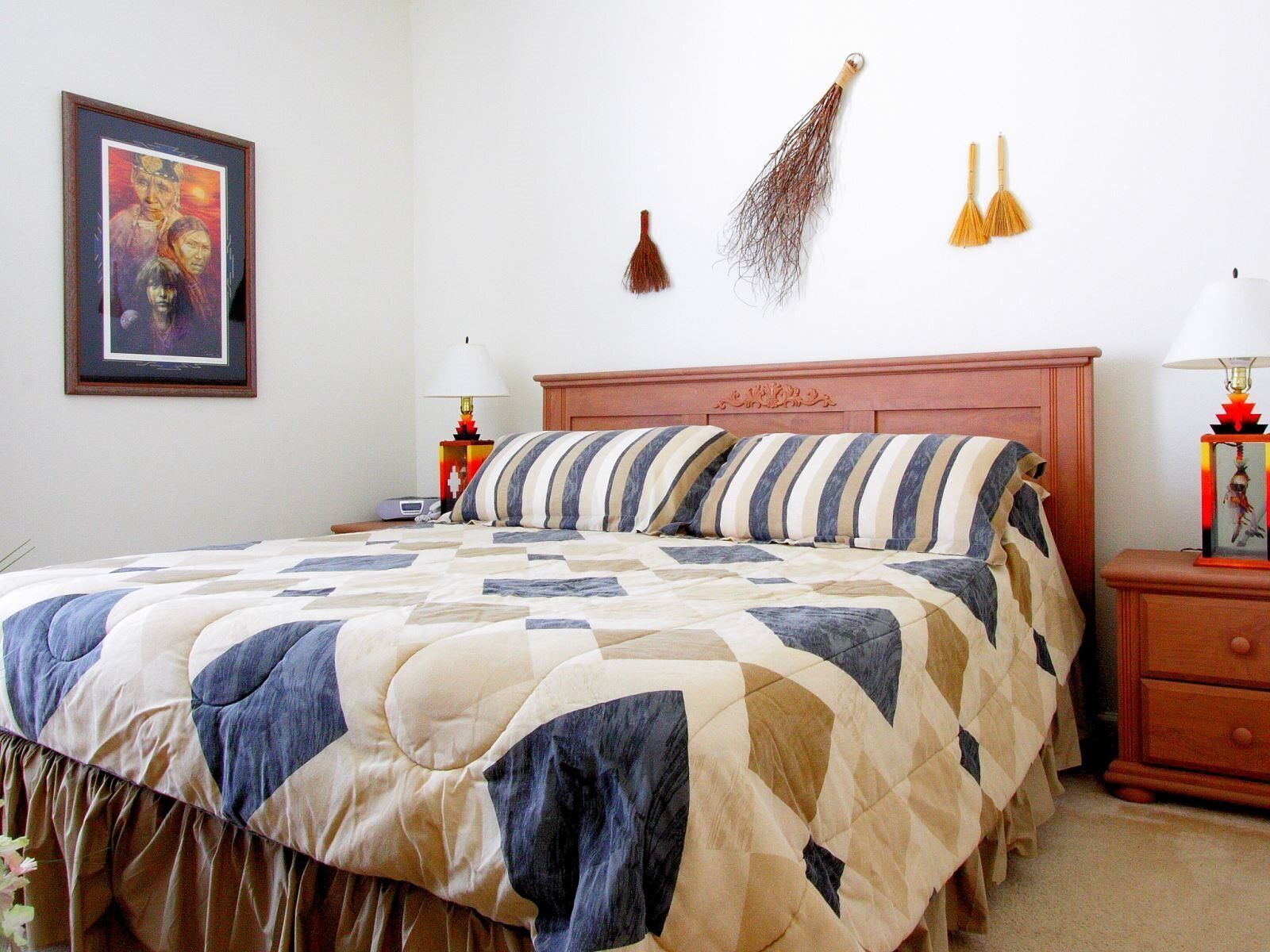 Une chambre à coucher avec un grand lit, une tête de lit en bois, deux tables de chevet avec des lampes, une lampe fixée au mur et un tableau encadré.