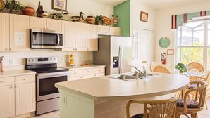 A kitchen with white cabinets, a microwave, a refrigerator, a stove, and a sink. There are wicker chairs and a small dining table. - 4 Bed 2269 (Kissimmee)