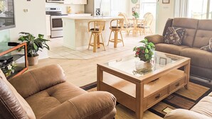 A modern living room with a brown leather sofa, a glass coffee table, and a brown armchair. A dining area with wicker chairs and a small island is visible in the background. - 4 Bed 2269 (Kissimmee)