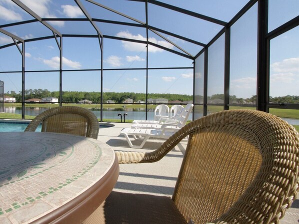 A covered outdoor area with a pool, wicker chairs, and a view of a lake and buildings.