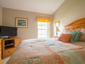 Ein Schlafzimmer mit einem Bett, einem Fernseher auf einem Holzboden, einem Fenster mit Vorhängen und einem Bild an der Wand.