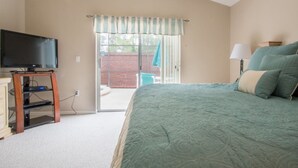 Un dormitorio con cama, televisor sobre un soporte de madera y puerta corrediza de vidrio que da a un espacio exterior.