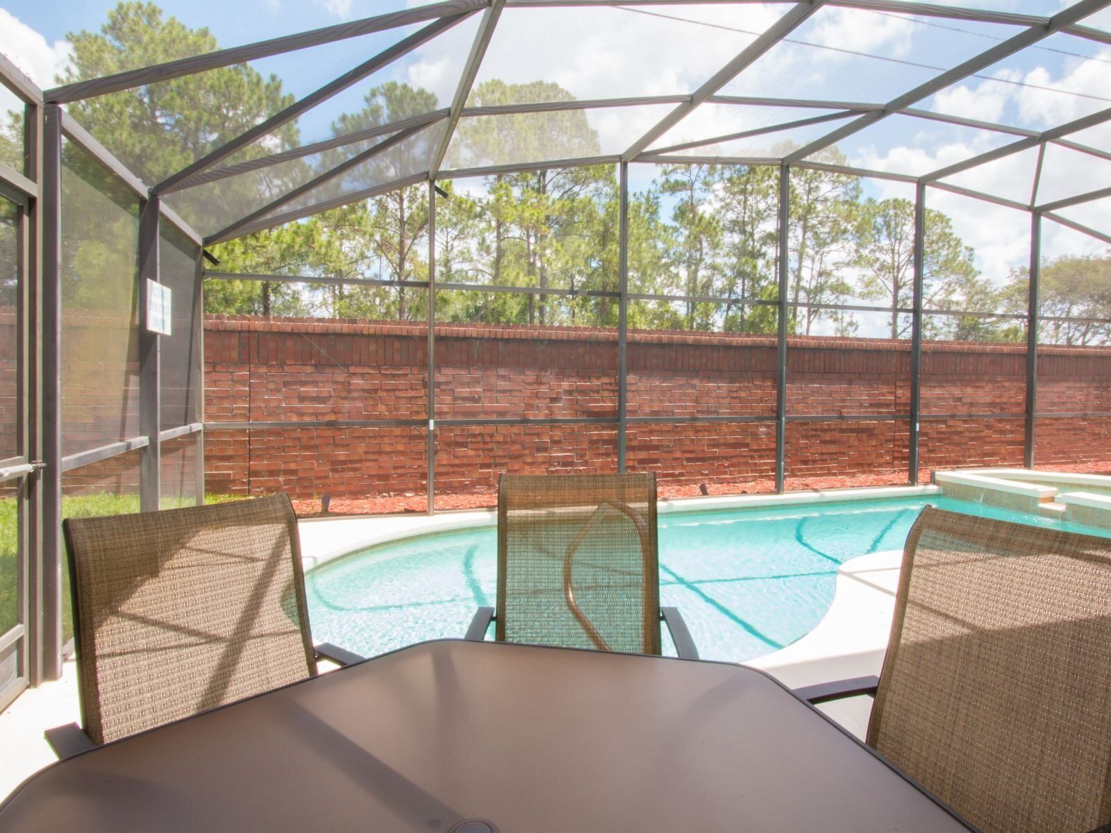A covered outdoor area with a pool, wicker furniture, and a brick wall in the background.