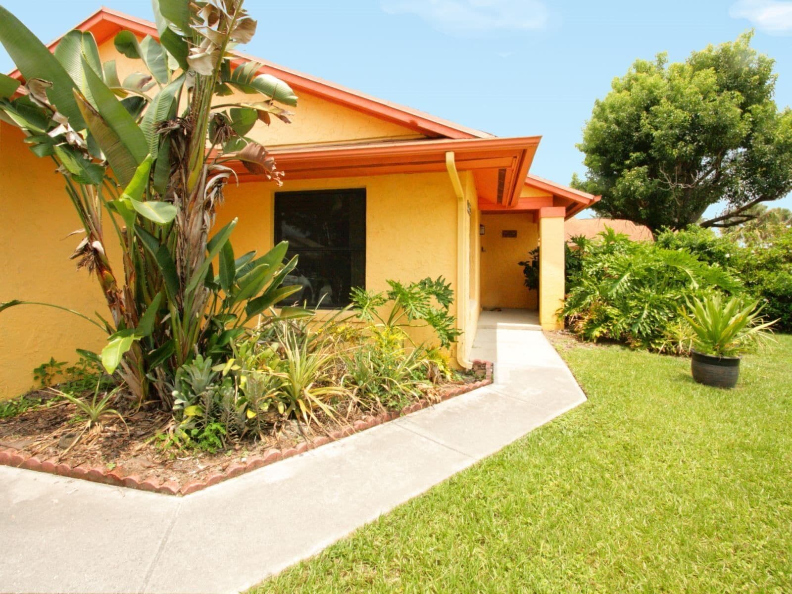 A yellow building with a red roof, surrounded by a well-maintained garden with various plants and a paved walkway.