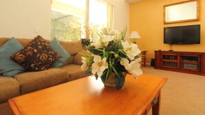 A living room with a brown sofa, a wooden coffee table, a vase with white flowers, a television, and a window with a view of trees. - 6 Bed 2525 (Kissimmee)
