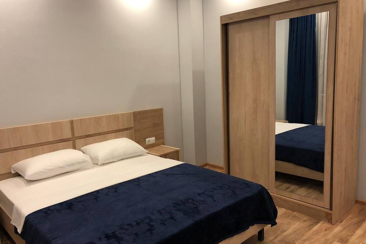 Una cama bien hecha con una colcha azul oscuro y dos almohadas blancas, un cabecero de madera, una mesita de noche con una lámpara y un mueble de espejo grande con cortina azul.