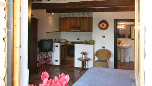 Une pièce rustique comprenant un lit, une chaise, une petite table et un coin cuisine visible à travers une porte ouverte.