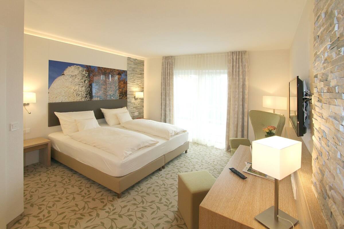 A hotel room with a large bed, a desk with a lamp, a chair, and a TV mounted on the wall.