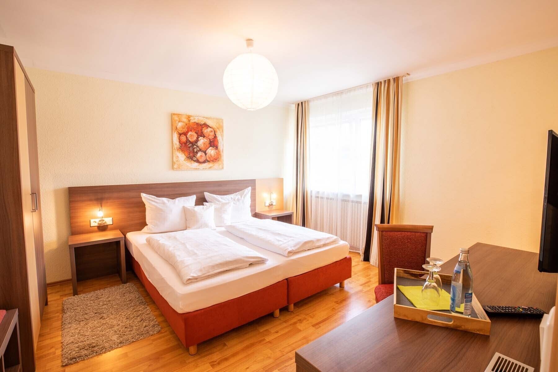 Camera d'albergo con un letto matrimoniale, comodini con lampade, un quadro appeso al muro, una finestra con tende, una scrivania in legno con un vassoio e una televisione.