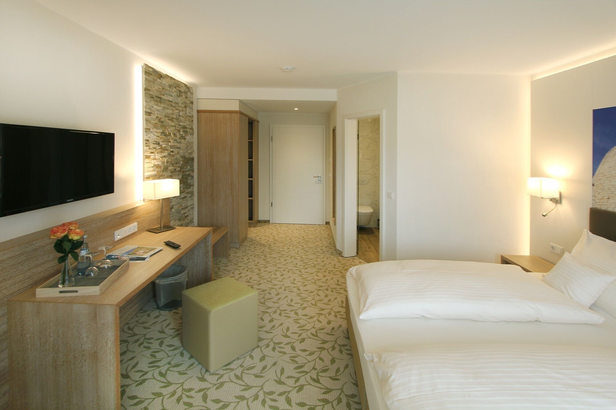 A hotel room with a bed, a desk with a lamp, a television, and a bathroom visible through an open door.