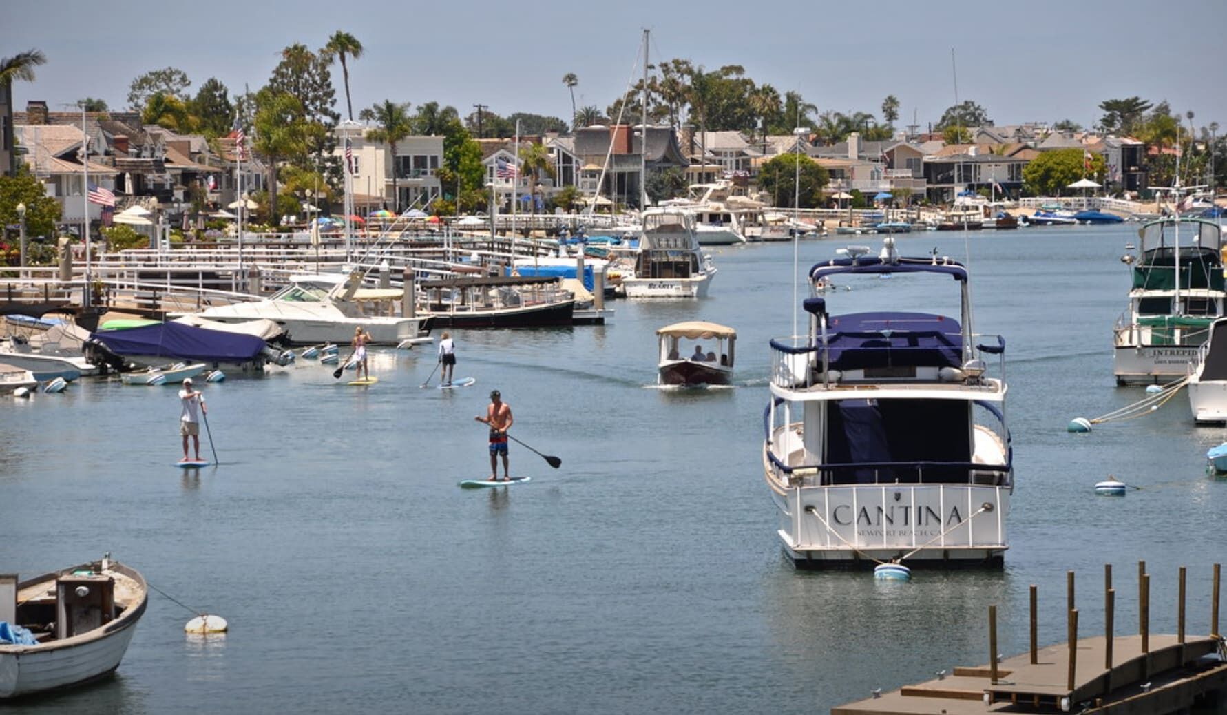 A marina with boats docked, a paddleboarder, and a sailboat named 'CANTINA'.
