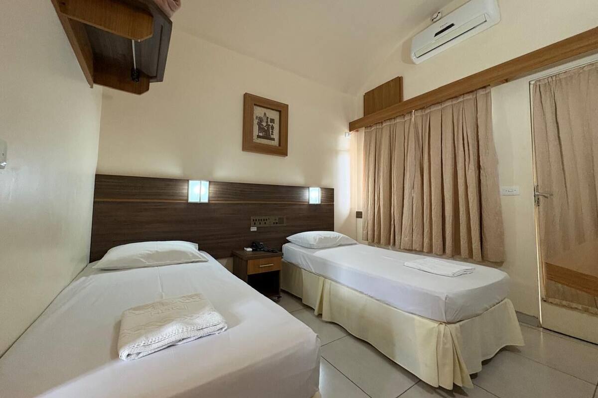 Habitación de hotel con dos camas individuales, cabecera de madera, mesita de noche, un cuadro enmarco, unidad de aire acondicionado y una puerta con cortinas.