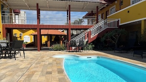 A swimming pool with a tiled deck, outdoor seating, and a covered area with stairs leading to a balcony. - Kanoa Hotel (Santo Antônio da Platina)