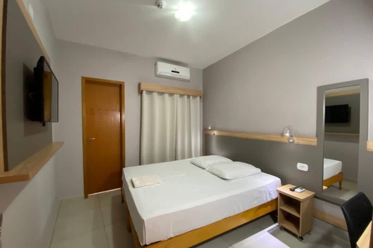 Un dormitorio con cama, mesita de noche, espejo y televisor instalado en la pared.