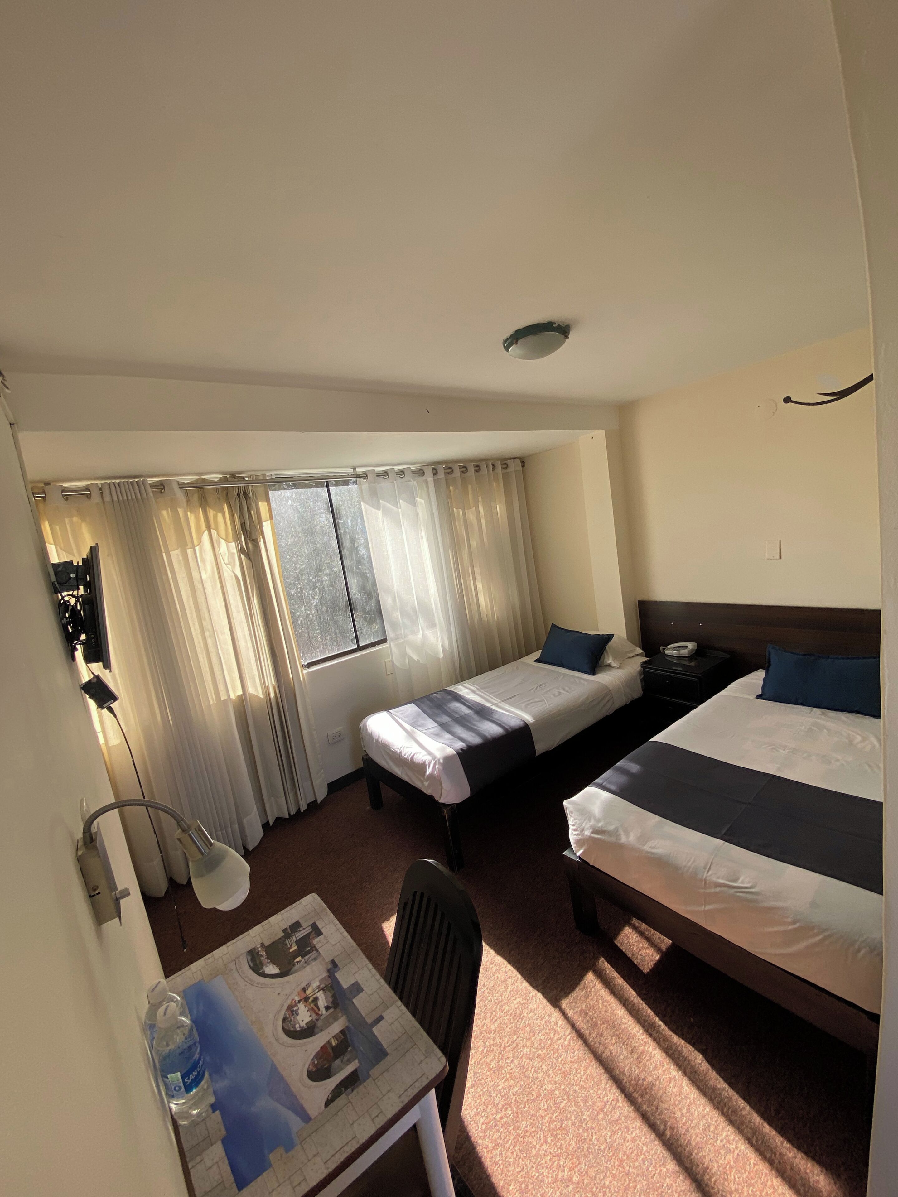 A hotel room with two beds, a desk, a chair, a table with a water bottle, and a window with curtains.