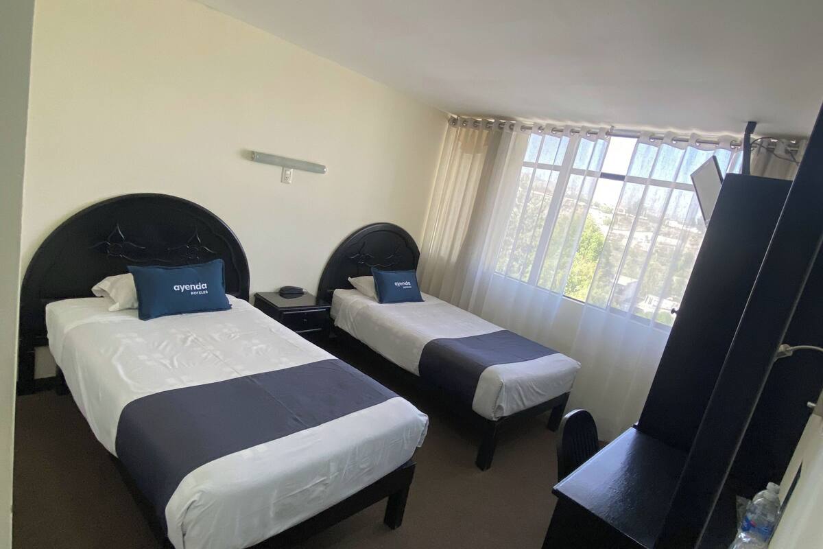 Une chambre d’hôtel avec deux lits, un bureau, une chaise, une armoire et une fenêtre avec des rideaux.