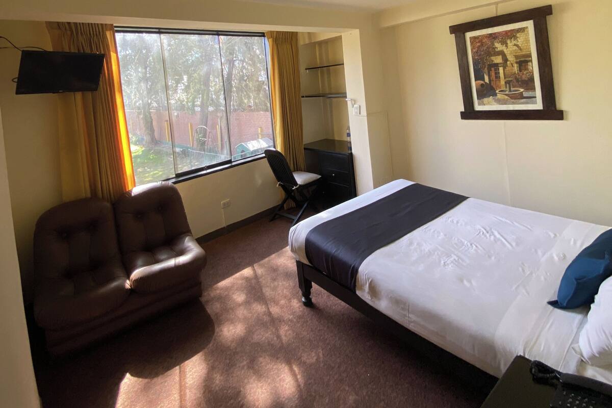 Une chambre d’hôtel avec un lit, un bureau, une chaise, une fenêtre avec des rideaux, un tableau au mur et un luminaire au plafond.