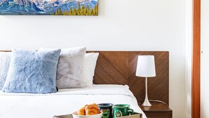 A neatly made bed with a wooden headboard, a bedside table with a lamp, and a tray with croissants and coffee mugs. - Mountain Luxe (Canmore)