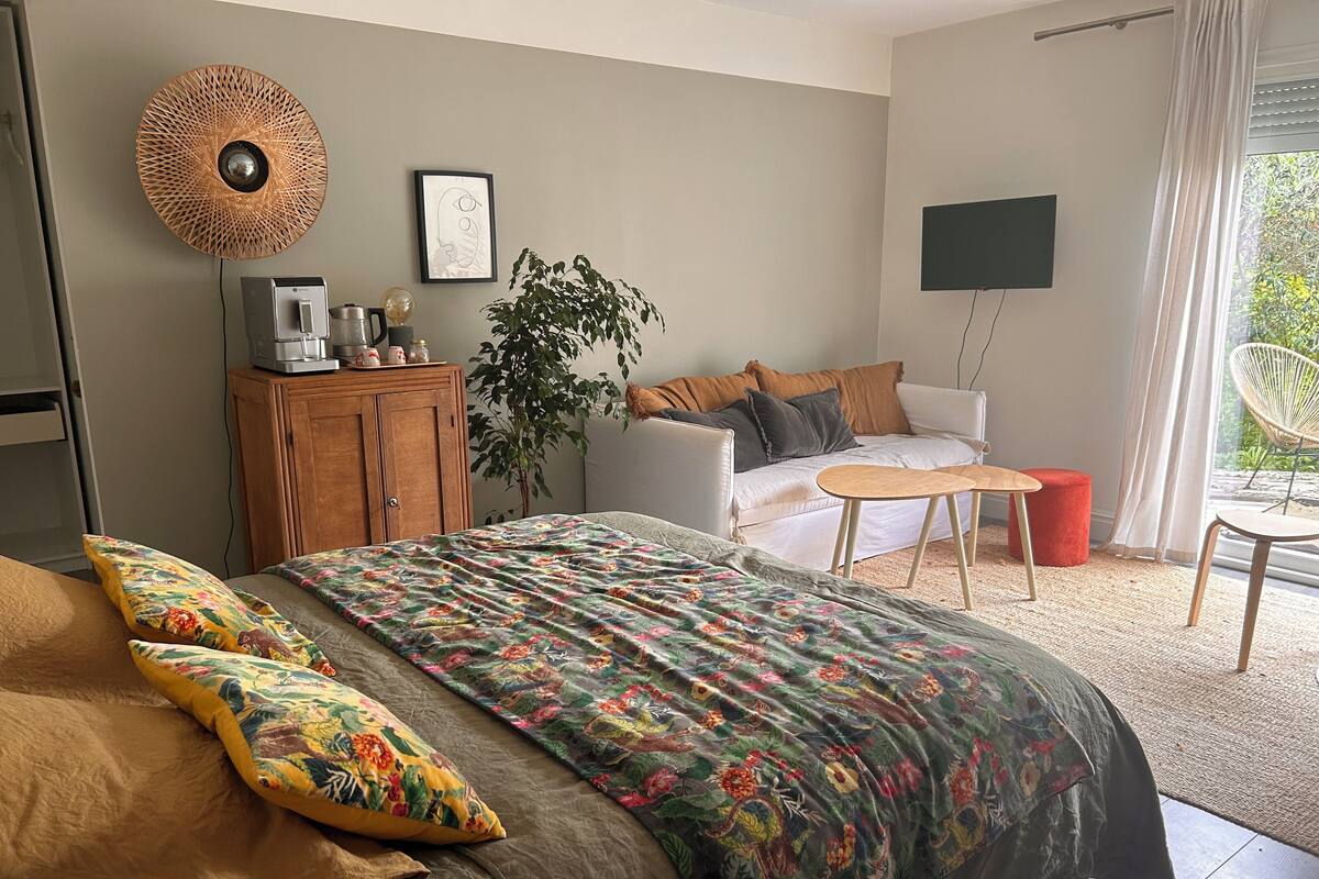 A bedroom with a bed, a sofa, a small table, a TV, a plant, and a window with a view.