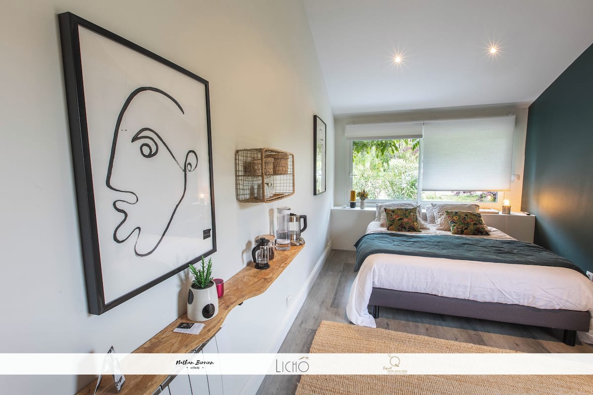 A modern bedroom with a large bed, a wooden bedside table with a coffee maker, a framed abstract artwork, and a window with a view of greenery.