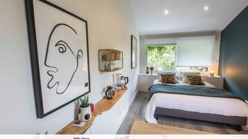 A modern bedroom with a large bed, a wooden bedside table with a coffee maker, a framed abstract artwork, and a window with a view of greenery.