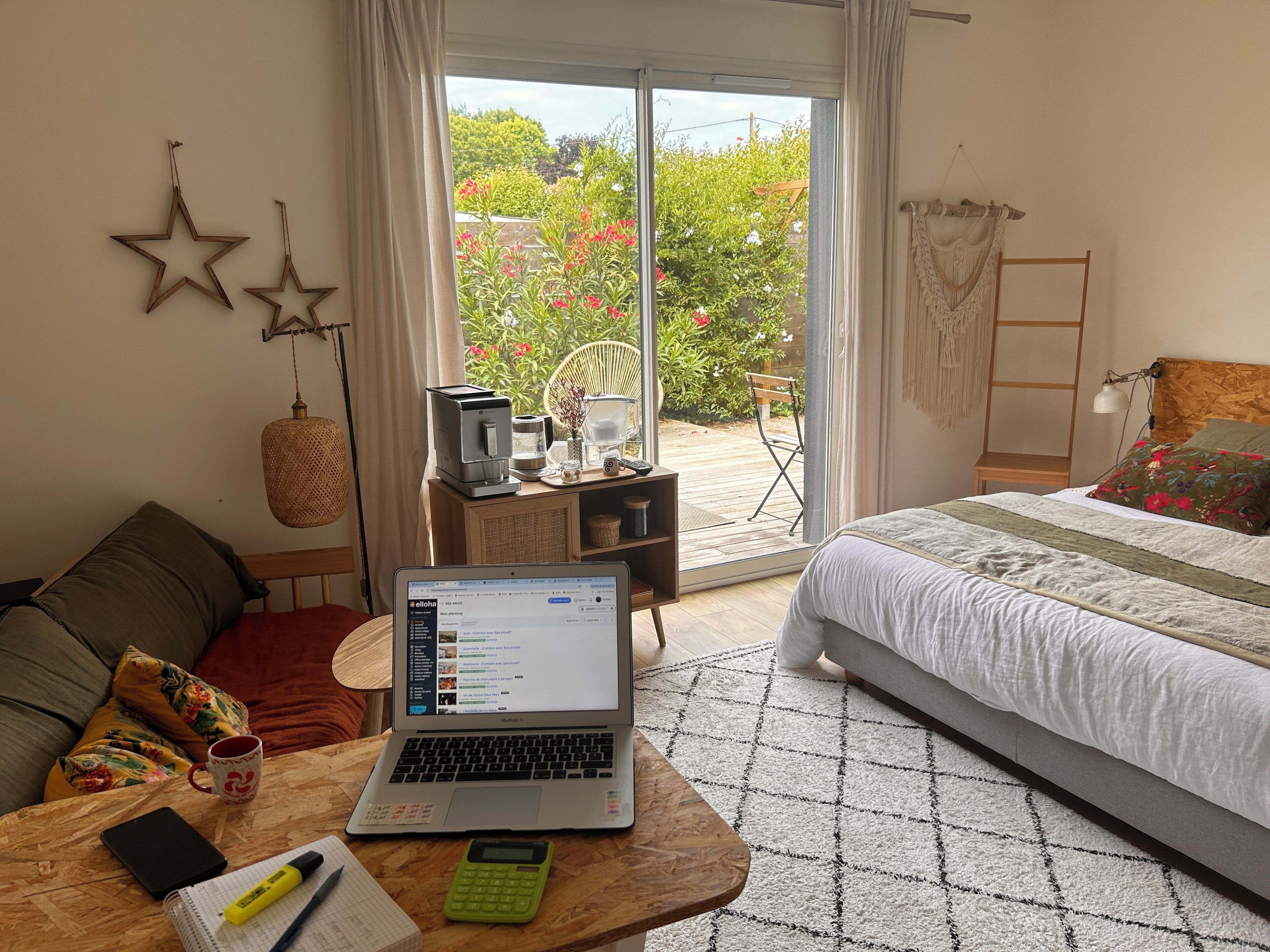 A bedroom with a laptop on a wooden table, a bed with a white duvet, and a view of a garden through a sliding door.