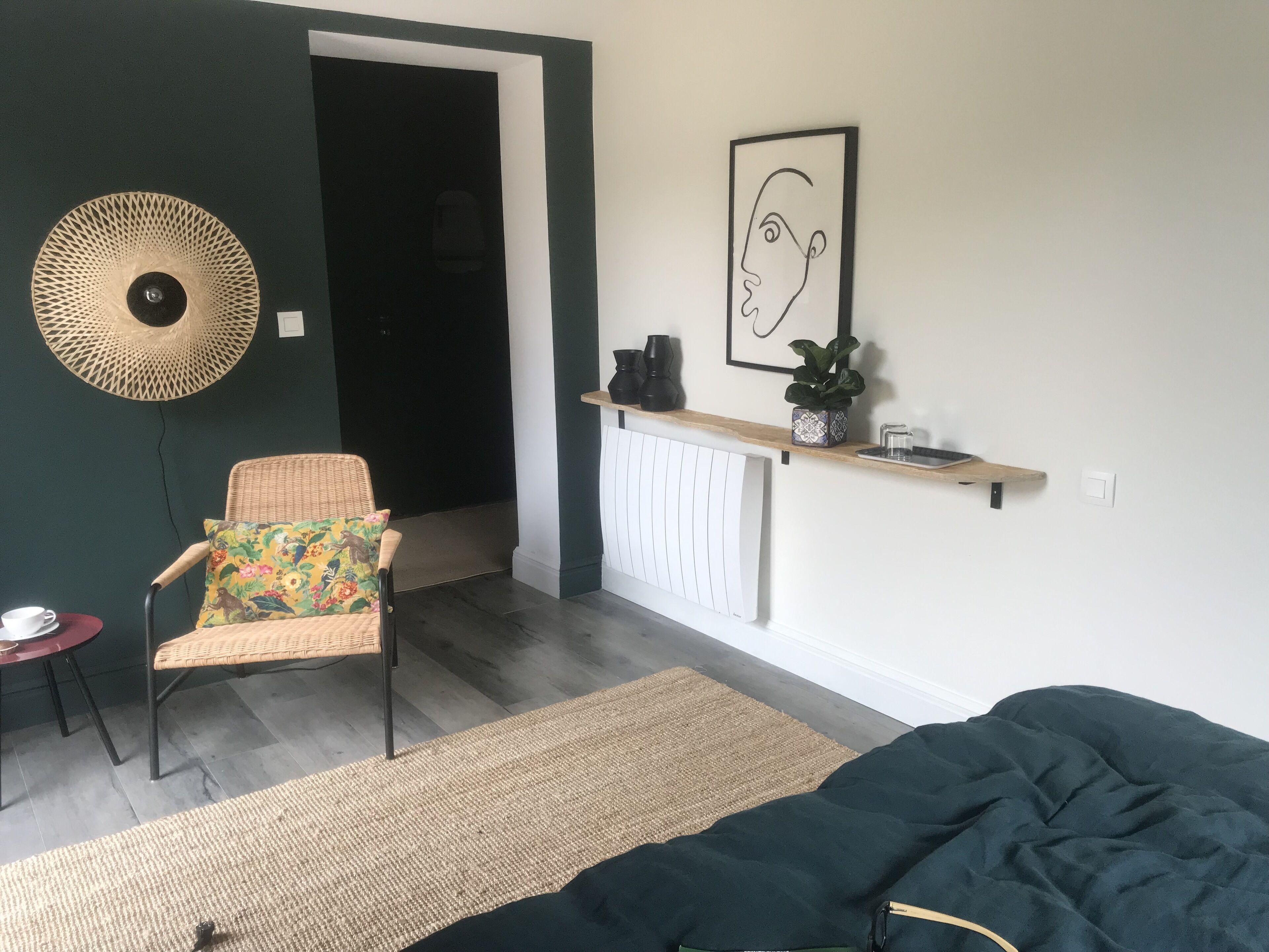 A room with a wicker chair, a radiator, a shelf with a plant, and a framed artwork on the wall.