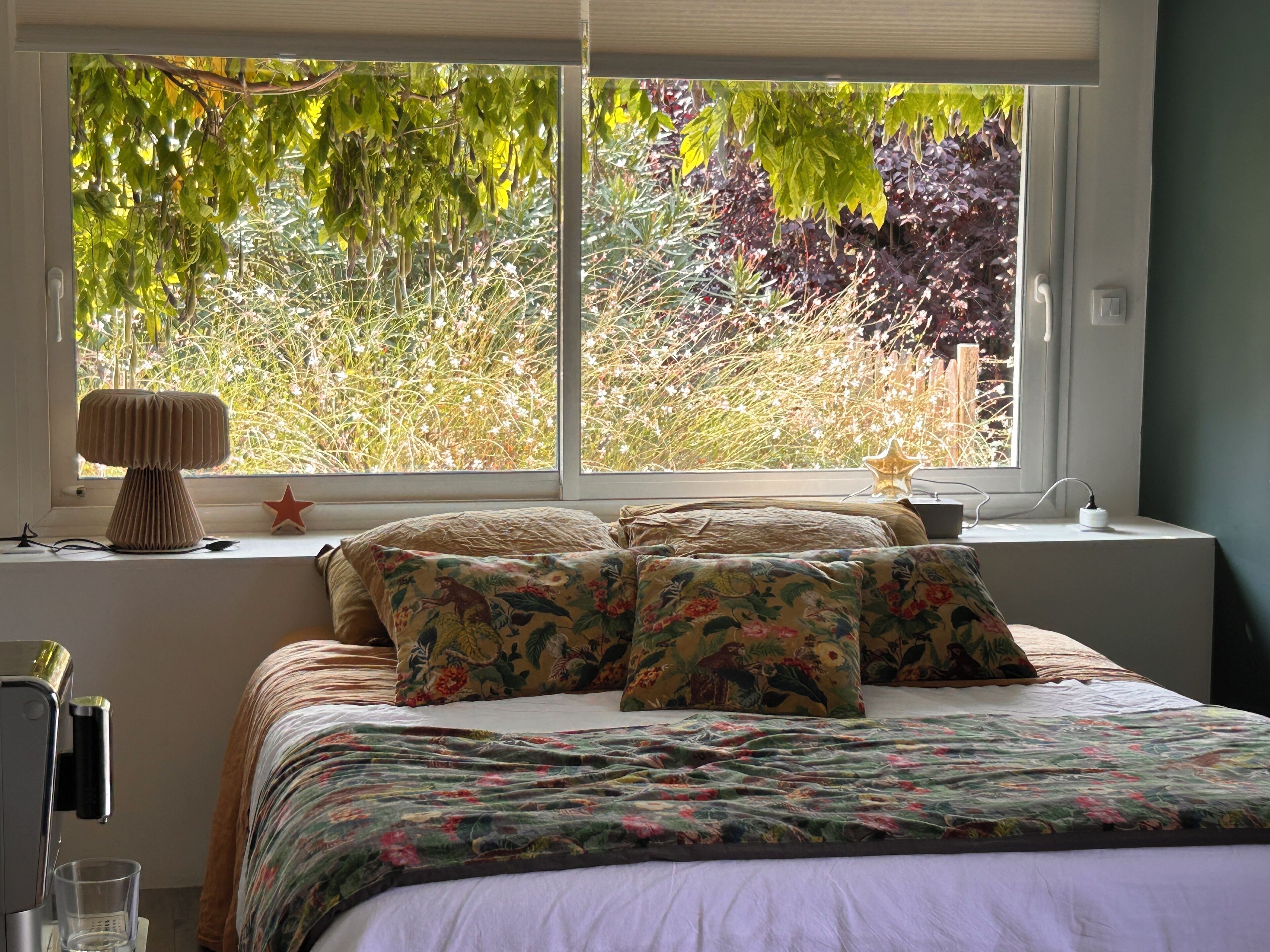 Un dormitorio con cama, ropa de cama floral, una lámpara de noche y una ventana con vistas a árboles y plantas.