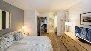 Quarto de hotel com cama, televisão sobre uma cômoda de madeira, espelho e porta que dá para outro aposento.