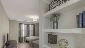 Une chambre avec deux lits, une télévision, une bibliothèque remplie de livres et d’objets décoratifs, et une fenêtre avec des rideaux.