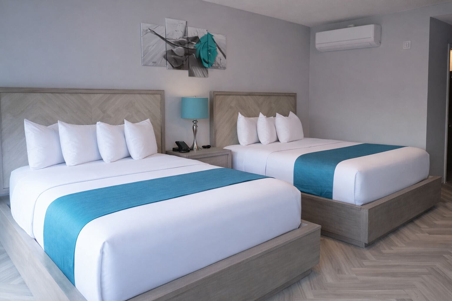 Ein Hotelzimmer mit zwei Betten, einem Nachttisch mit Lampe, einer Wand-Klimaanlage und einem dekorativen Gegenstand über den Betten.