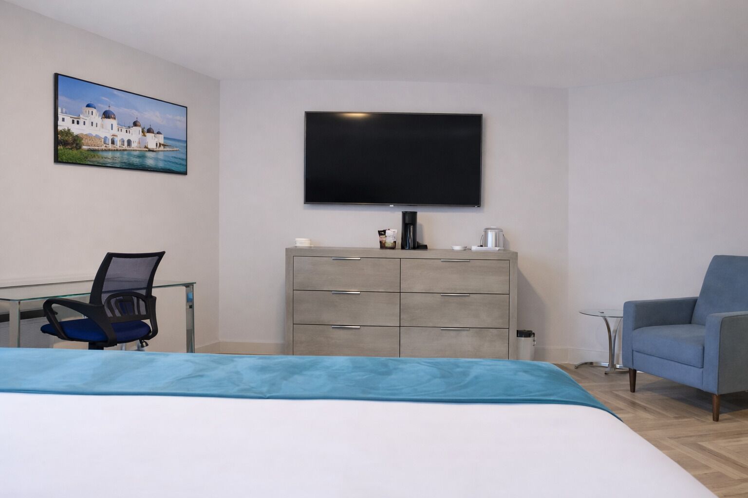 Quarto de hotel com cama, escrivaninha, cadeira, cômoda, televisão e um quadro na parede.