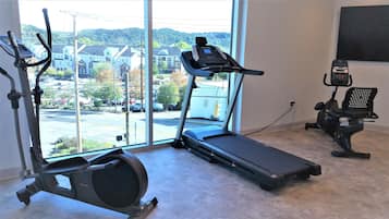A home gym with a treadmill, elliptical, and stationary bike, with a view of houses and a parking lot through large windows.