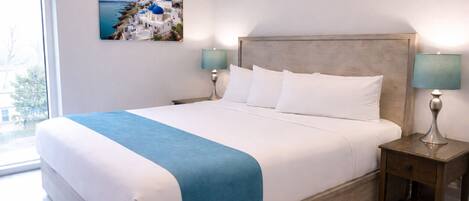 A neatly made bed with a blue accent, two bedside lamps, a picture of a coastal village, and a wooden nightstand.