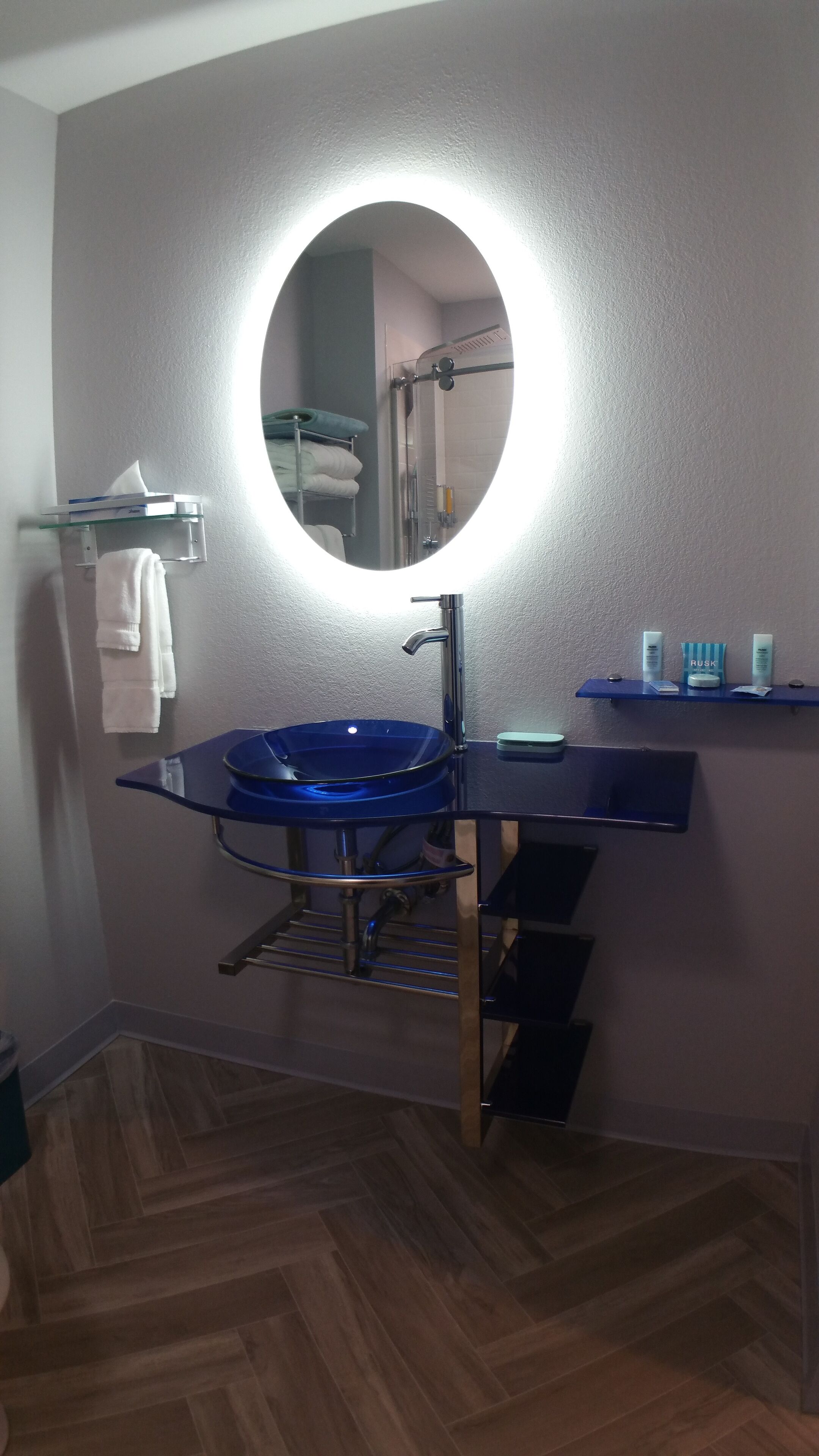 Een badkamer met een blauwe wastafel, een ronde spiegel met verlichting, een handdoekrek en een plank met toiletartikelen.