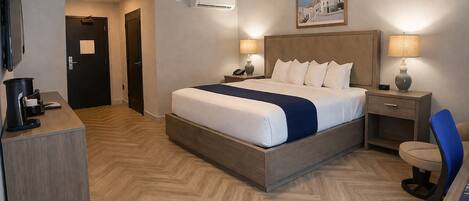 A hotel room with a bed, bedside tables, a desk, a chair, a coffee machine, and an air conditioning unit.