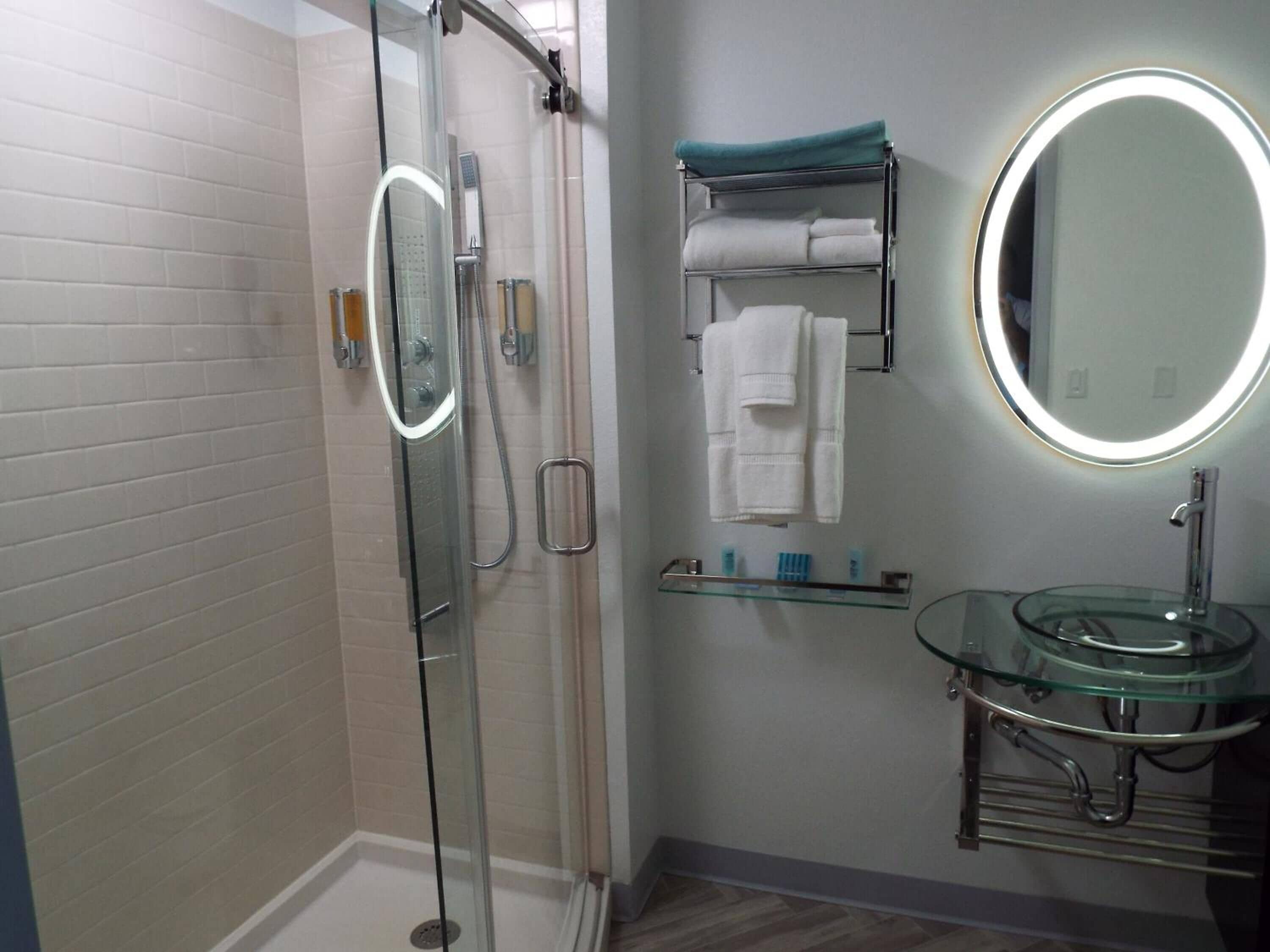 A modern bathroom with a glass shower enclosure, a glass sink with a round mirror, and a towel rack with folded towels.