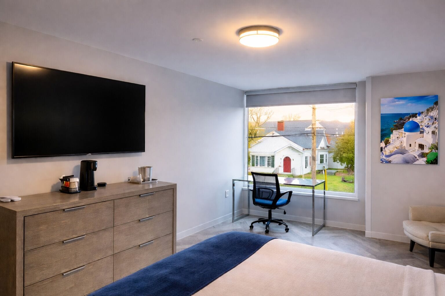 Camera d'albergo moderna con una grande TV, una scrivania con sedia, un letto e una finestra vista su una casa bianca e cielo blu.