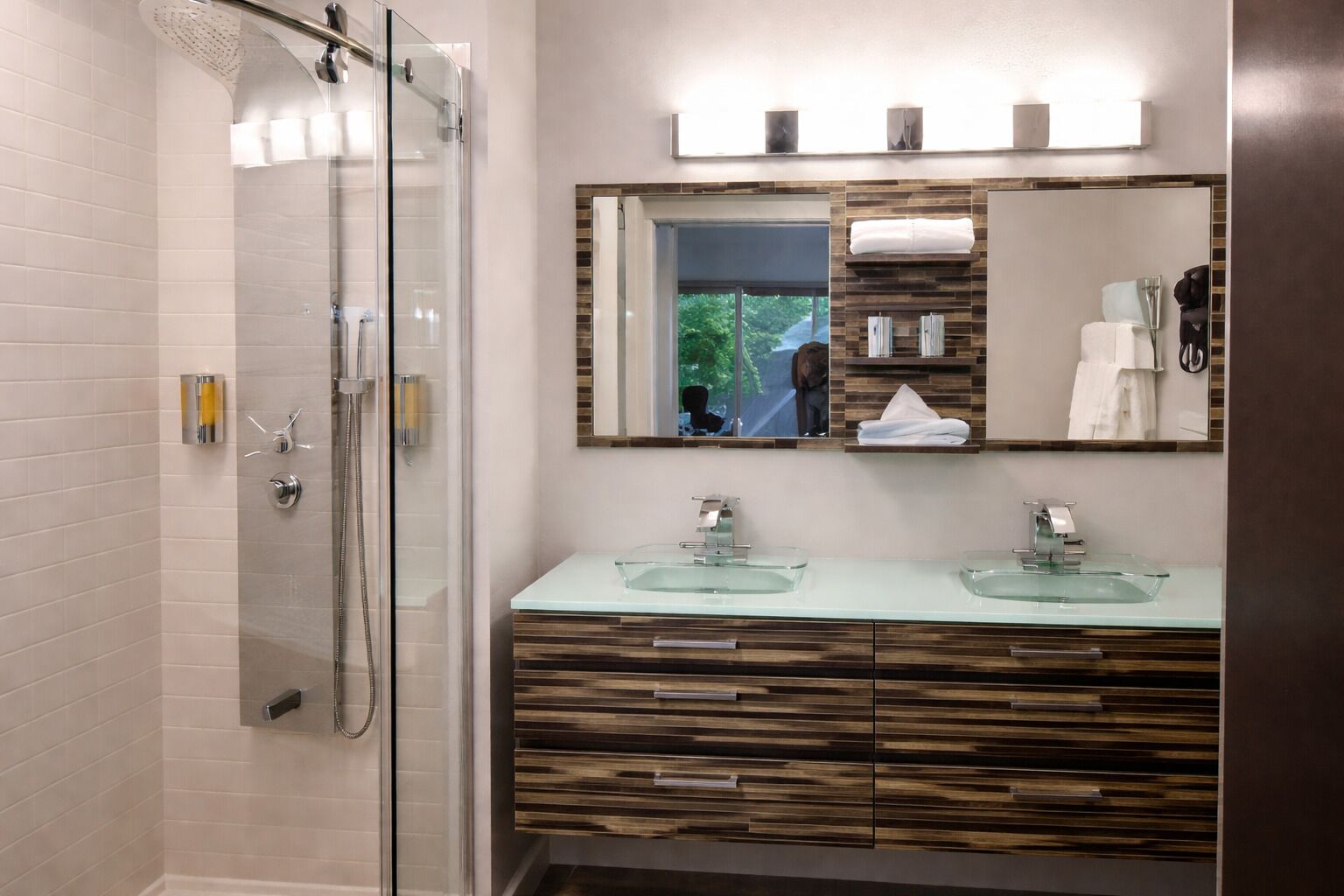 A modern bathroom with a glass-enclosed shower, two glass sinks, and a large mirror with a view of the outdoors.