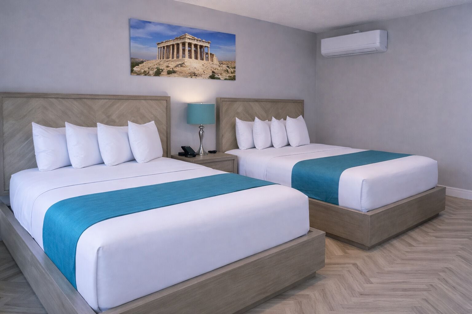 A hotel room with two beds, a nightstand with a lamp, a wall-mounted air conditioner, and a picture of the Parthenon.