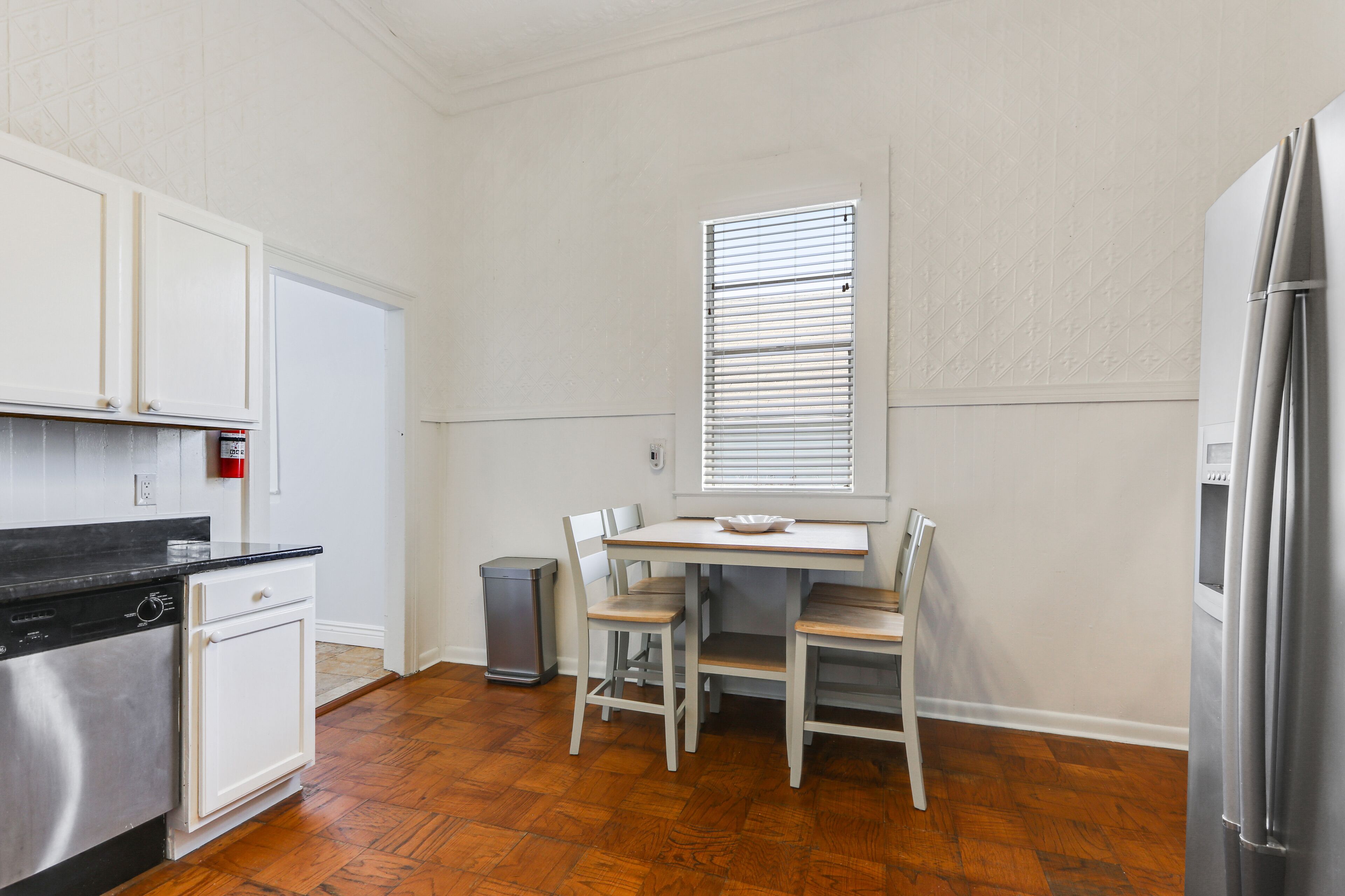 A compact kitchen with white cabinets, a black countertop, a stainless steel refrigerator, and a window with blinds.