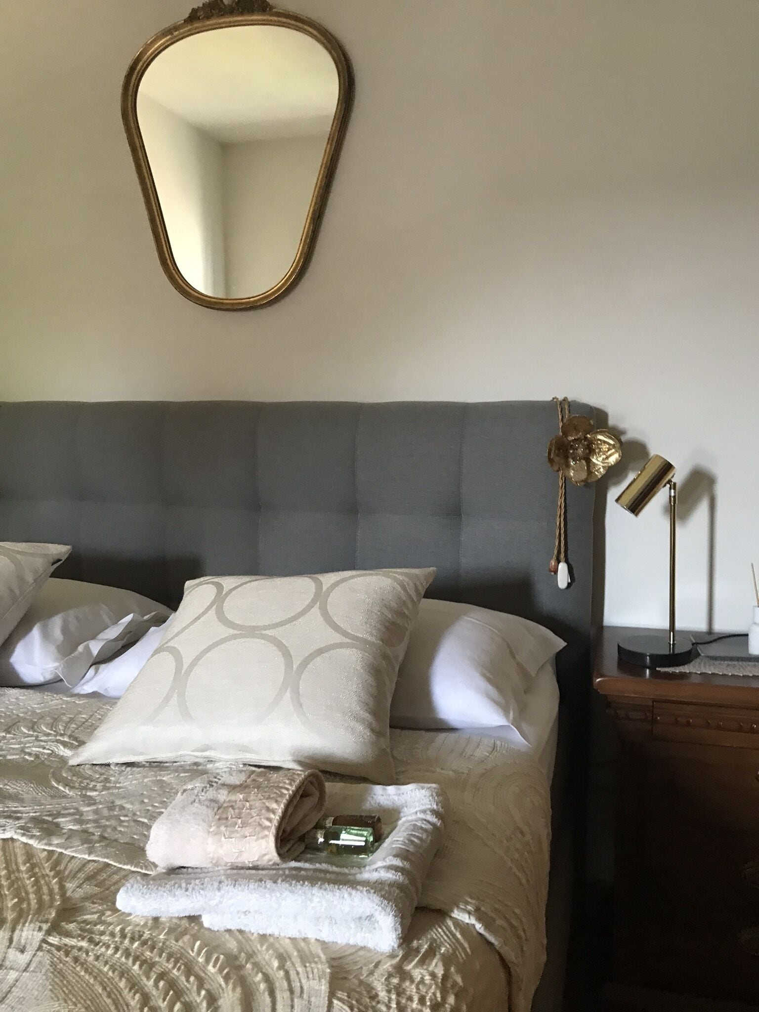 A neatly made bed with a grey headboard, a mirror above it, and a bedside table with a lamp and a small decorative item.
