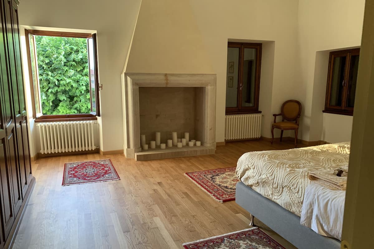 Quarto com teto inclinado, lareira, cama, cadeira e janela com vista para o verde.
