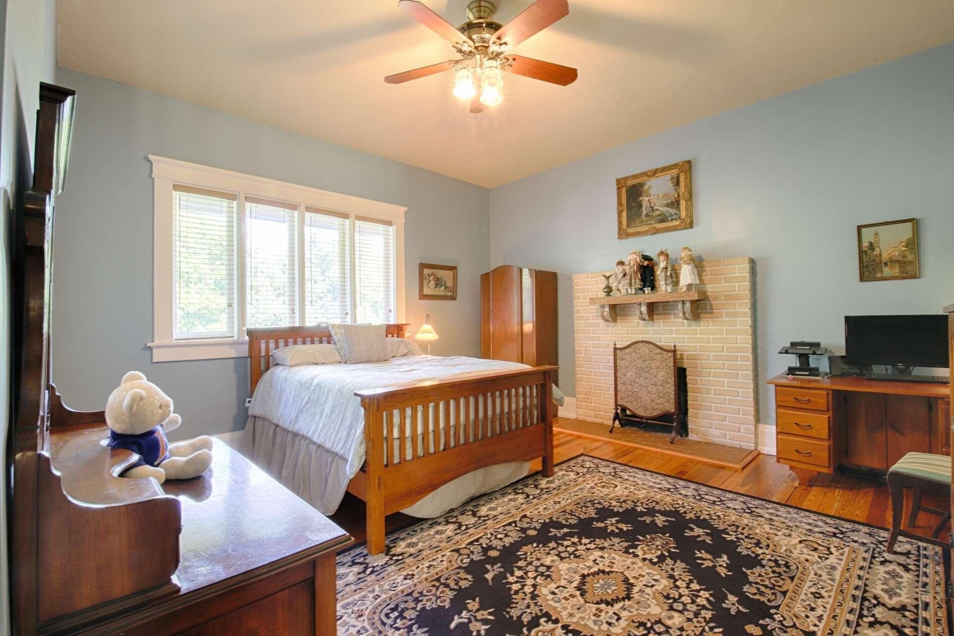 A bedroom with a bed, a dresser, a desk, a chair, a fireplace, and a ceiling fan.