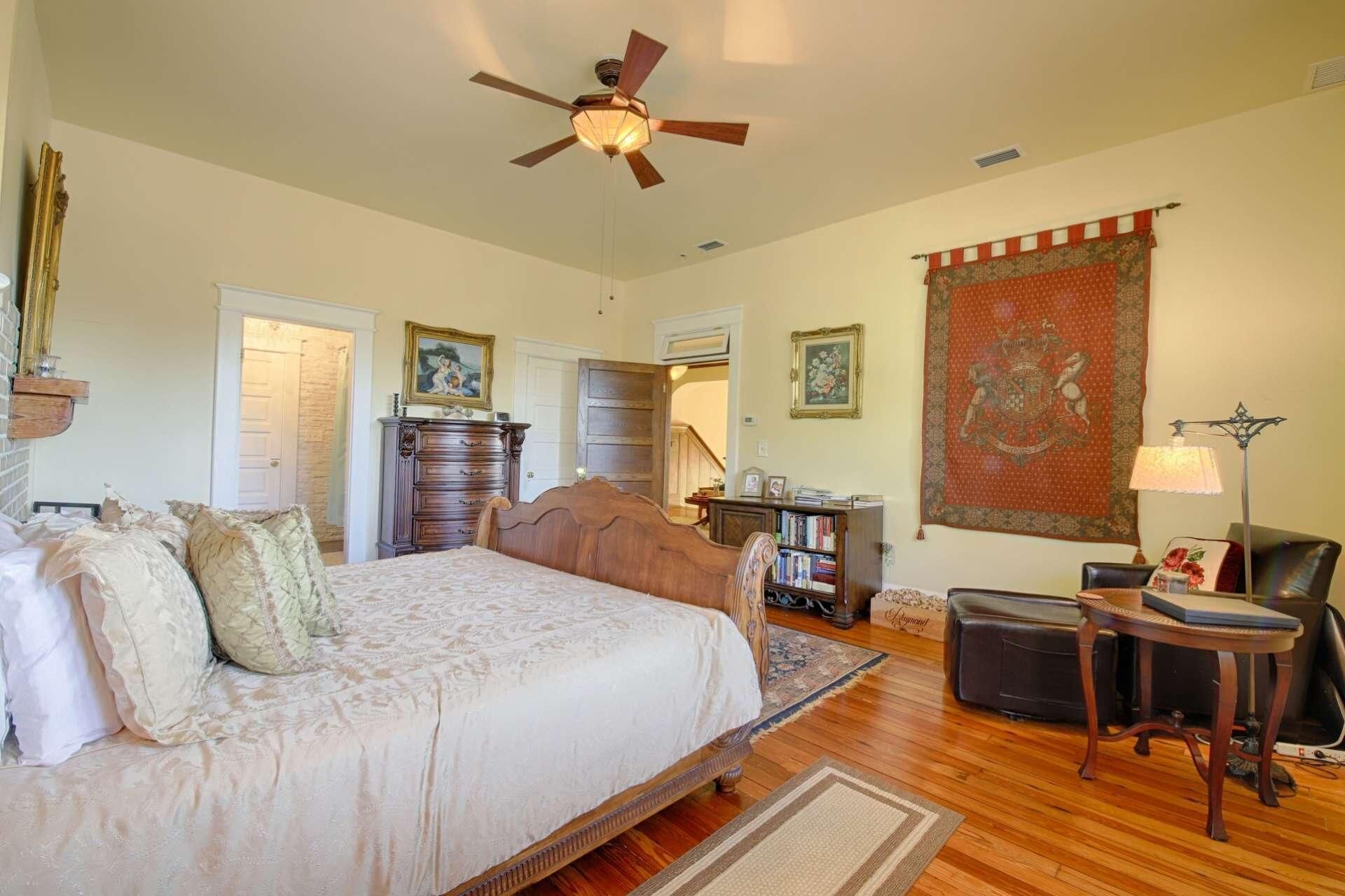 A spacious bedroom with a large bed, a wooden dresser, a desk, and a chair. There is a ceiling fan, a tapestry, and a bookshelf.