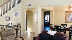 A living room with a brown leather sofa, patterned armchair, and a coffee table. There is a staircase leading to another level, and a dining area in the background.