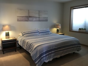 Un dormitorio con cama, dos mesitas de noche, una lámpara de noche, ventana con vistas y una pared decorada con obras de arte.