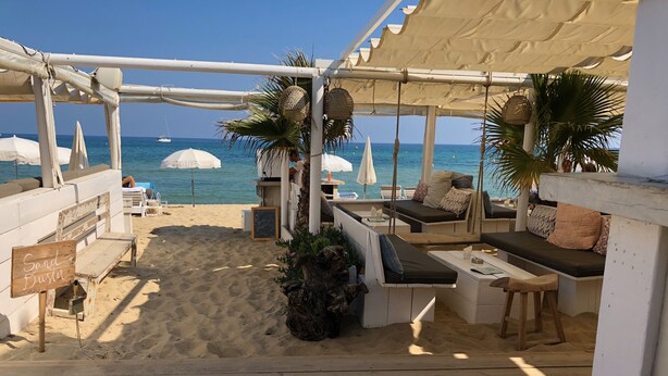 A beachside seating area with wooden benches and tables, a sign reading "Sand Buster," and a view of the ocean. - Atmospheric villa with nice sea view in a quiet green area (La Croix-Valmer)