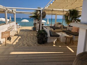 A beachside seating area with wooden benches and tables, a sign reading "Sand Buster," and a view of the ocean. - Atmospheric villa with nice sea view in a quiet green area (La Croix-Valmer)